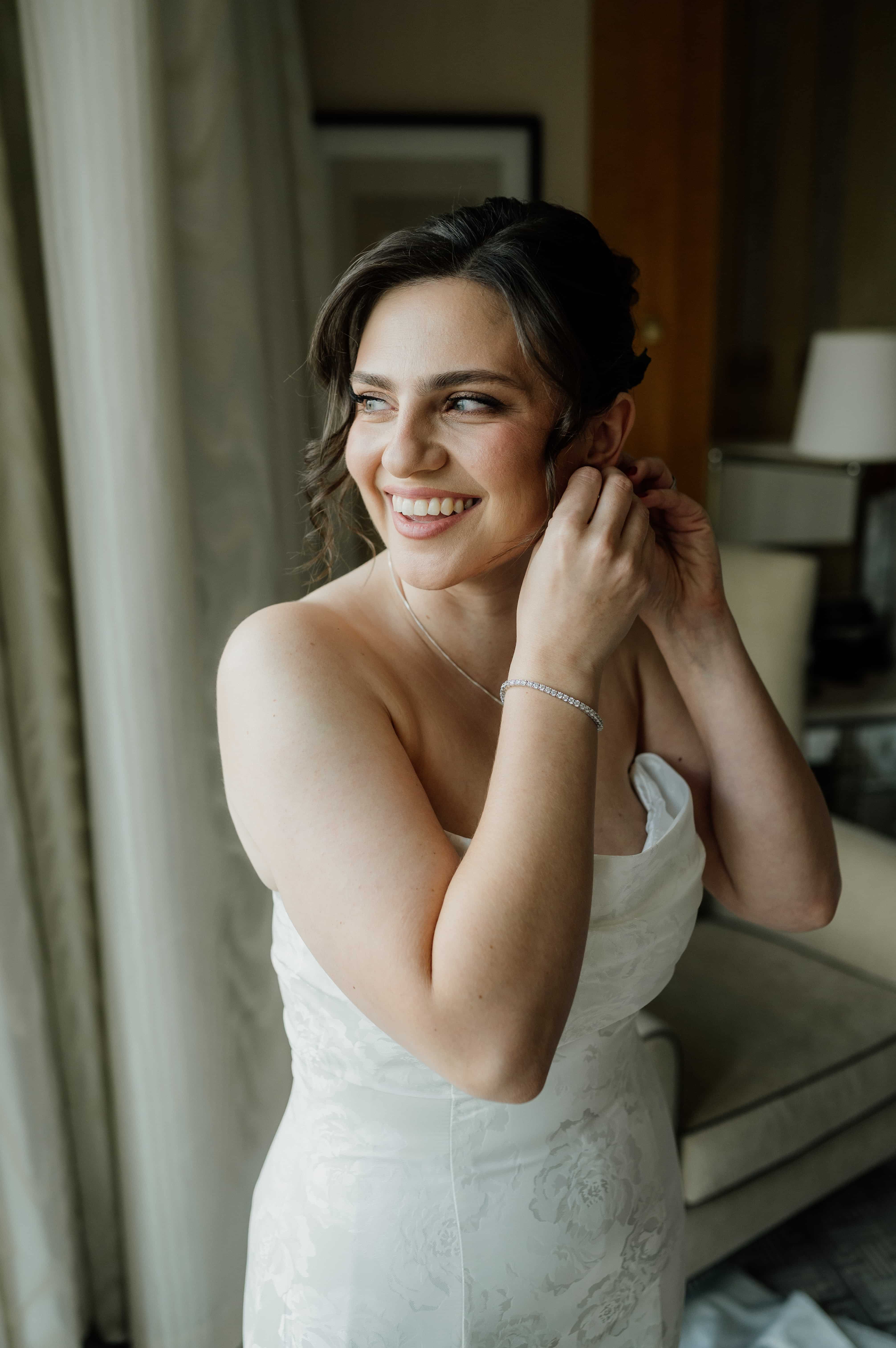 Smiling bride putting on earring in soft window light